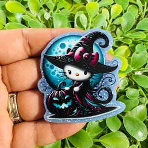 May include: A die-cut sticker featuring a white cat character wearing a black witch hat with a pink bow, standing next to a black jack-o'-lantern. The background shows a large blue moon with swirling decorative patterns and glittery accents.