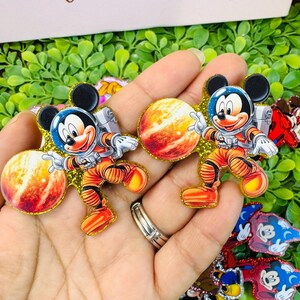 K&ouml;nnte beinhalten: Zwei Mickey Mouse Pins zum Thema Astronaut. Jeder Pin zeigt Mickey in einem orangefarbenen Raumanzug mit blauem Helm, silbernem Rucksack und einem Planeten. Die Pins sind mit goldenem Glitzer umrandet und vor einem gr&uuml;nen Hintergrund platziert.
