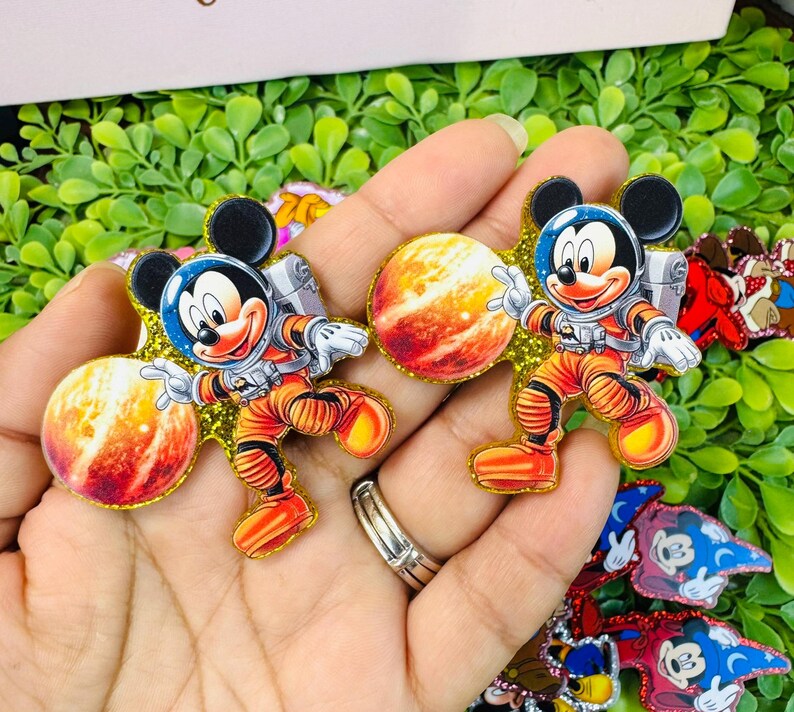 K&ouml;nnte beinhalten: Zwei Mickey Mouse Pins zum Thema Astronaut. Jeder Pin zeigt Mickey in einem orangefarbenen Raumanzug mit blauem Helm, silbernem Rucksack und einem Planeten. Die Pins sind mit goldenem Glitzer umrandet und vor einem gr&uuml;nen Hintergrund platziert.