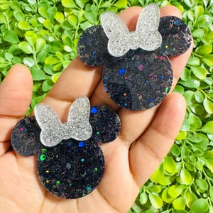 May include: Two glittery black Minnie Mouse-shaped accessories with silver bows. The black resin is embedded with colorful glitter. The accessories are held in a hand against a background of green leaves.