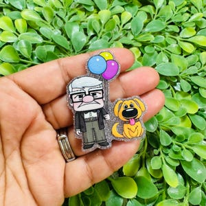 May include: A glittery acrylic pin featuring a cartoon elderly man with white hair and glasses wearing a suit, holding three colorful balloons in blue, yellow, and purple, standing next to a golden-yellow cartoon dog with a pink tongue.