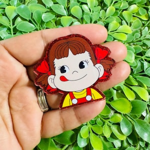 May include: A glittery acrylic pin featuring a cartoon character with brown hair, red bows, and a yellow shirt with red overalls. The character has a light skin tone, large black eyes, and a tongue sticking out to the side of the mouth.