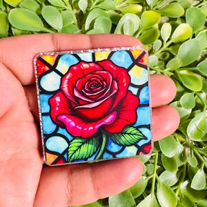 May include: Square-shaped decorative item featuring a vibrant red rose with green leaves against a stained-glass window design. The artwork has a detailed, colorful appearance with a silver glitter border.