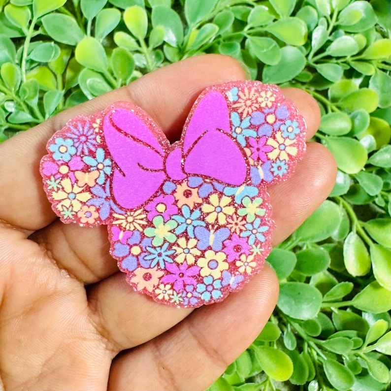 May include: A pink glittery Minnie Mouse-shaped accessory with a large bow. The ears and bow are solid pink, while the head is filled with colorful flowers in shades of blue, yellow, and pink. The accessory is held in a hand.