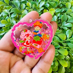 May include: A pink heart-shaped charm featuring Strawberry Shortcake and a friend. The charm has a clear center filled with pink glitter and star-shaped confetti. The characters are dressed in colorful outfits, with a strawberry theme.