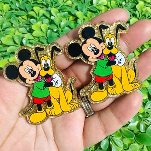 May include: Two glittery acrylic charms featuring Mickey Mouse and Pluto hugging. Mickey is in a green shirt and red shorts, Pluto is yellow. The charms have a gold glitter border and black accents.