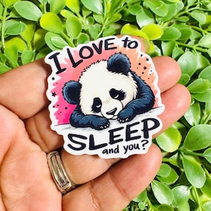 May include: A die-cut sticker featuring a sleepy panda resting its head on its paws. The background is a soft pink and orange gradient with small black stars. The text reads I LOVE TO SLEEP and you? in bold black lettering around the panda.