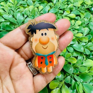 May include: A flat, glittery acrylic badge featuring the cartoon character Fred Flintstone. The character has black hair, a tan face, a large nose, and wears an orange tunic with black triangular spots and a blue necktie, all outlined in gold glitter.