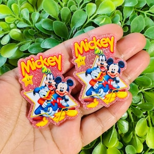 May include: Two pink glittery Mickey Mouse-themed embellishments. Each features Mickey, Donald, and Goofy on a star-shaped background. The word "Mickey" is written in yellow above the characters. The background is a lush green.