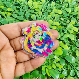 May include: A clear acrylic pin featuring a chibi-style illustration of a blonde character with blue eyes, wearing a blue and red outfit with a gold tiara. The design includes pink hearts and red roses against a dark blue circular background.