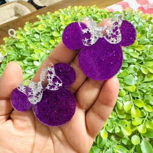 Pode incluir: Dois acess&oacute;rios roxos com glitter de resina em forma de cabe&ccedil;a da Minnie Mouse com la&ccedil;os transparentes com recortes de estrelas. Os acess&oacute;rios s&atilde;o segurados em uma m&atilde;o, com folhagem verde ao fundo. O glitter d&aacute; aos acess&oacute;rios uma apar&ecirc;ncia brilhante.