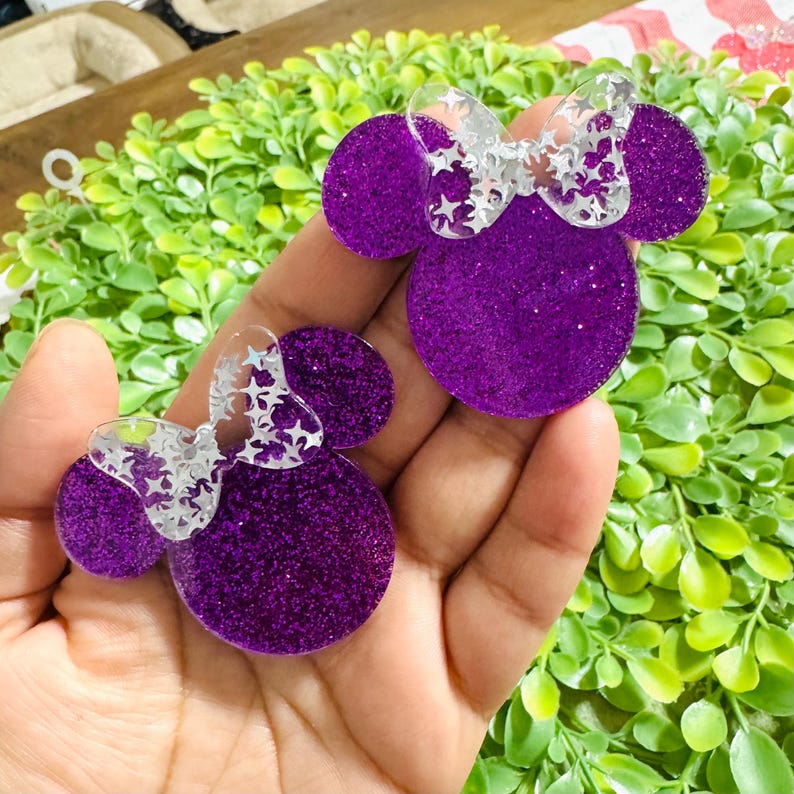 Pode incluir: Dois acess&oacute;rios roxos com glitter de resina em forma de cabe&ccedil;a da Minnie Mouse com la&ccedil;os transparentes com recortes de estrelas. Os acess&oacute;rios s&atilde;o segurados em uma m&atilde;o, com folhagem verde ao fundo. O glitter d&aacute; aos acess&oacute;rios uma apar&ecirc;ncia brilhante.