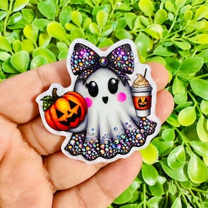 May include: A ghost-shaped acrylic charm featuring a black bow covered in multicolored rhinestones, a bright orange jack-o'-lantern pumpkin, and a coffee cup with a pumpkin face graphic. The base of the ghost is decorated with colorful rhinestones.