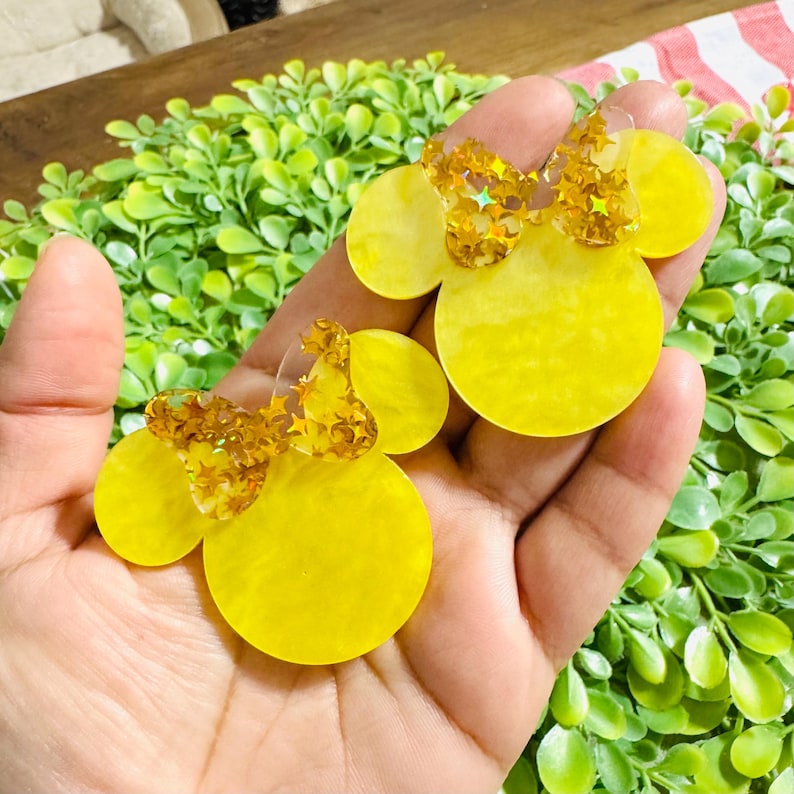 May include: Two yellow Minnie Mouse-shaped earrings with gold glitter bows. The earrings are held in a hand against a background of green foliage. The earrings are made of a translucent yellow material.