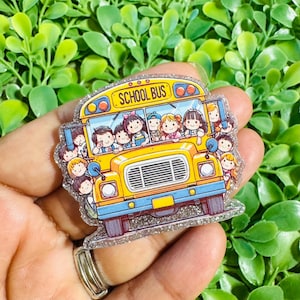 May include: A yellow school bus pin with a glittery finish featuring a cartoon illustration of multiple children looking out the windows. The bus has a front grille, headlights, and the text SCHOOL BUS printed in black on the top front panel.