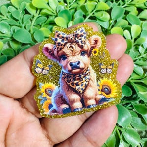 May include: A glittery, die-cut acrylic charm featuring a fluffy Highland calf wearing a leopard print bow and bandana. The calf is surrounded by yellow sunflowers and small butterflies against a sparkling gold background with a textured finish.