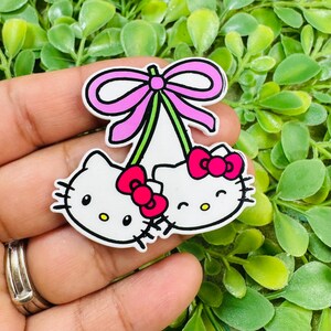 May include: A white enamel pin featuring two Hello Kitty character heads joined by green stems topped with a large pink bow. Each character has a small bright pink bow on its ear, black whiskers, black eyes, and a yellow nose, all outlined in black.