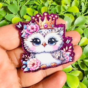 May include: A glittery purple owl-shaped embellishment with a gold crown and floral accents. The owl has large, expressive eyes and is surrounded by pink flowers and butterflies. The design is detailed and colorful.