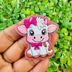 May include: A glittery pink and white cow-shaped embellishment with a pink bow on its head and a pink bow tie. The cow has brown spots and is smiling. The edges of the embellishment are outlined in pink glitter.