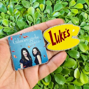 May include: Two square and oval-shaped pins with glittery edges. One pin features the Gilmore Girls: A Year in the Life logo with two women, and the other is a yellow oval with the red text Luke's, shaped like a coffee cup silhouette.