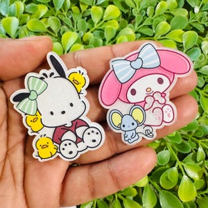 May include: Two cartoon character acrylic charms. One features a white dog with a green bow, surrounded by yellow chicks. The other is a pink bunny with a blue bow, holding a blue mouse. Both charms have a clear, glossy finish.