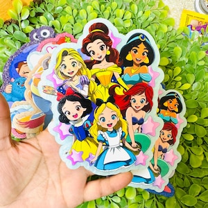 May include: A collection of colorful stickers featuring various Disney princesses. The stickers showcase characters like Belle, Jasmine, Ariel, Snow White, and Alice, with a star-shaped border. The stickers are held in a hand against a backdrop of green foliage.