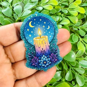May include: A glittery pin featuring a central candle with a yellow flame, set against a dark blue circular background with yellow crescent moons and stars. The base of the candle is surrounded by stylized blue and purple floral petals.