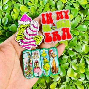 May include: Three decorative acrylic pins featuring holiday themes. One pin shows a green character in a pink hat holding a striped treat. Another is a glittery square with red and green text reading IN MY GRINCH ERA. The third is a rectangular pin with four panels depicting a Christmas tree, a girl with a gift, a green character, and a dog.