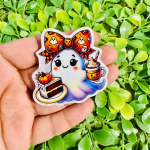 May include: A die-cut sticker featuring a white ghost with a smiling face, wearing a large orange and black bow patterned with ghosts and pumpkins. The ghost holds a slice of chocolate cake with a pumpkin topper and a mug with a jack-o'-lantern face.