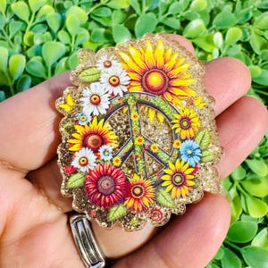 May include: A decorative badge featuring a peace sign surrounded by colorful sunflowers, daisies, and other flowers. The badge has a gold glitter border and is set against a green background.