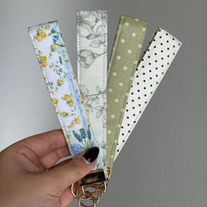 May include: Four fabric key fobs with gold-coloured hardware are held in a hand. The key fobs display diverse patterns: floral, botanical, polka dots, and a floral design. The key fobs are in shades of white, blue, yellow, green, and black.