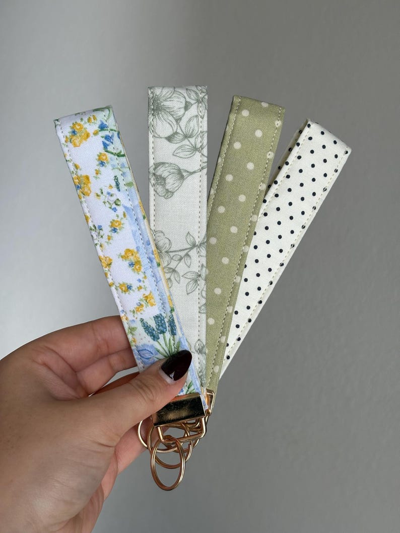 May include: Four fabric key fobs with gold-coloured hardware are held in a hand. The key fobs display diverse patterns: floral, botanical, polka dots, and a floral design. The key fobs are in shades of white, blue, yellow, green, and black.