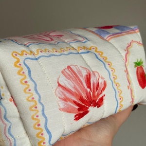 May include: A quilted, rectangular pouch with a white background and a repeating pattern of red seashells, strawberries, and decorative borders in blue, yellow, and pink. The pouch is held in a hand.
