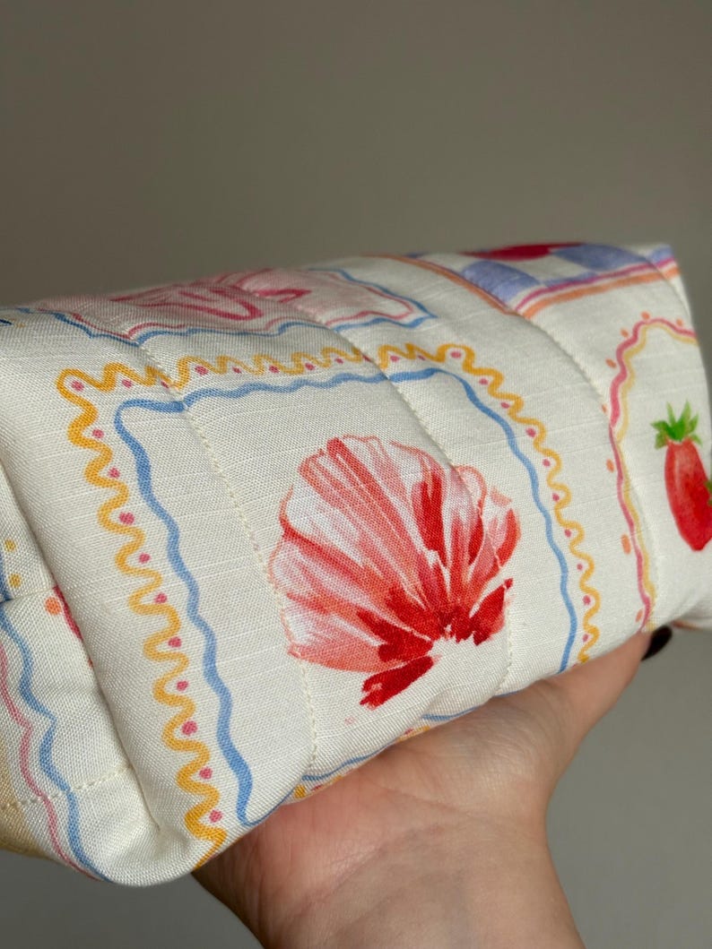 May include: A quilted, rectangular pouch with a white background and a repeating pattern of red seashells, strawberries, and decorative borders in blue, yellow, and pink. The pouch is held in a hand.