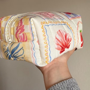 May include: A quilted cosmetic bag with a colorful floral and seashell pattern. The bag features a zipper closure and is held in a hand. The bag's design includes yellow, pink, blue, and red elements.