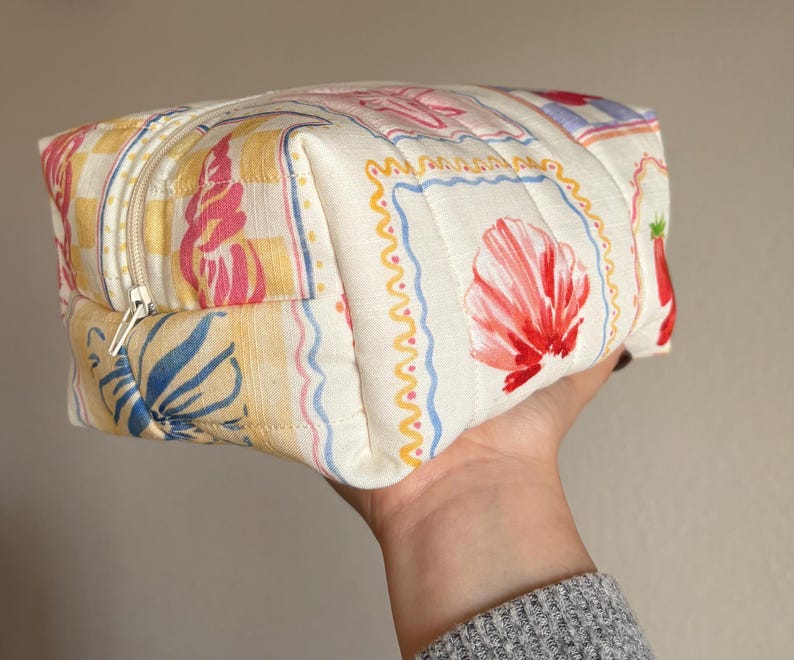 May include: A quilted cosmetic bag with a colorful floral and seashell pattern. The bag features a zipper closure and is held in a hand. The bag's design includes yellow, pink, blue, and red elements.
