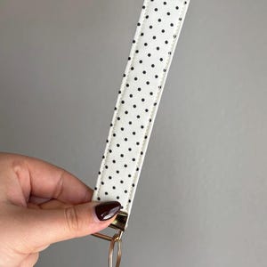 May include: A white key fob wrist strap featuring a black polka dot design. It has a gold-coloured metal ring and hardware. The wrist strap is held by a hand against a grey background.