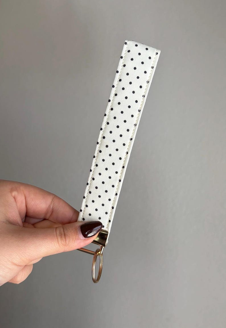 May include: A white key fob wrist strap featuring a black polka dot design. It has a gold-coloured metal ring and hardware. The wrist strap is held by a hand against a grey background.