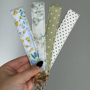 May include: Four fabric wrist keychains with gold-colored hardware. The patterns include a floral design with yellow and blue flowers, a gray floral pattern, a green polka dot pattern, and a white polka dot pattern. Each keychain is approximately 6 inches long.