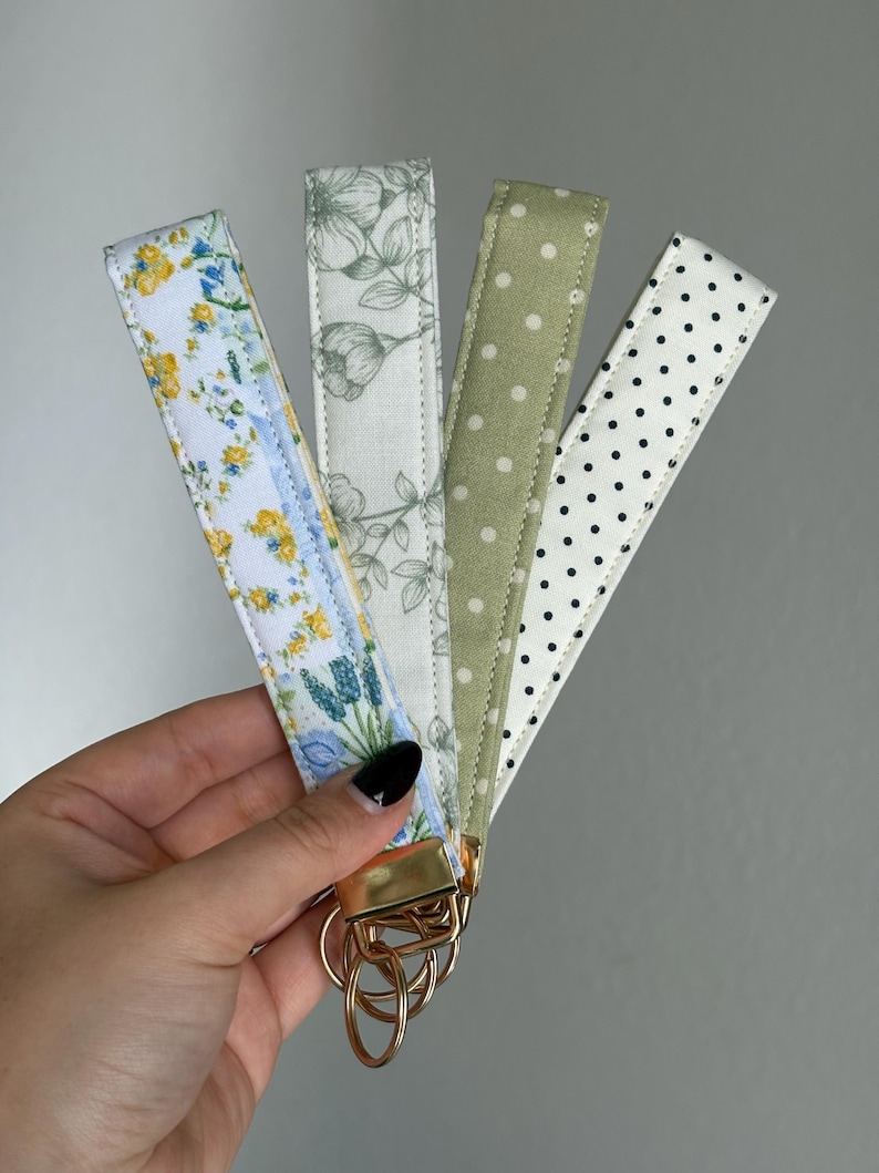 May include: Four fabric wrist keychains with gold-coloured hardware. The patterns include a floral design with yellow and blue flowers, a grey floral pattern, a green polka dot pattern, and a white polka dot pattern. Each keychain is approximately 15 cm long.