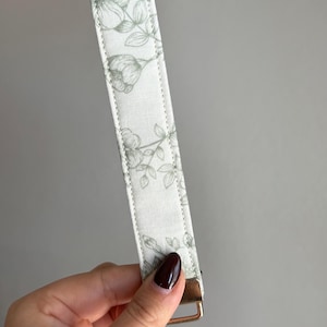 May include: A white key fob wristlet with a floral pattern in shades of green. The wristlet has a gold-coloured metal clasp and a loop for keys. The fabric is stitched along the edges.