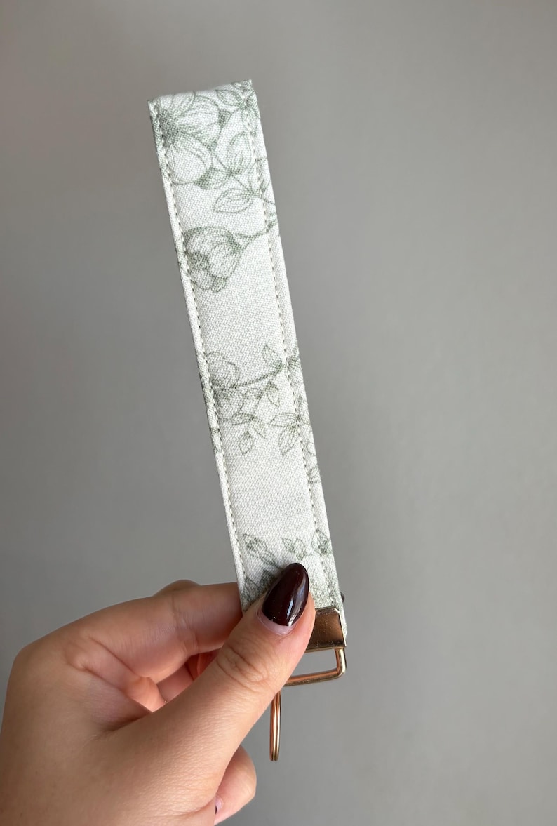 May include: A white key fob wristlet with a floral pattern in shades of green. The wristlet has a gold-coloured metal clasp and a loop for keys. The fabric is stitched along the edges.