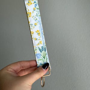 May include: A floral key fob featuring a white background with yellow and blue flowers. It has a gold-coloured metal clasp and ring. The fabric is patterned with small flowers and green leaves.