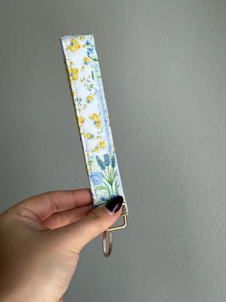 May include: A floral key fob featuring a white background with yellow and blue flowers. It has a gold-coloured metal clasp and ring. The fabric is patterned with small flowers and green leaves.