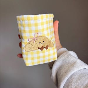 Può includere: Una piccola pochette rettangolare in tessuto con motivo a quadretti vichy gialli e bianchi. La parte anteriore è decorata con un ricamo raffigurante un croissant beige con un viso sorridente, piedini marroni e un fiocchetto rosa chiaro.