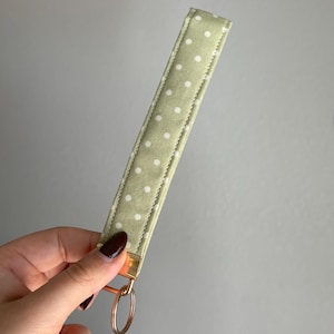 May include: A pale green key fob wrist strap with a white polka dot design. It features a gold-tone metal clasp and a copper-coloured key ring. The fabric has a soft, textured feel.