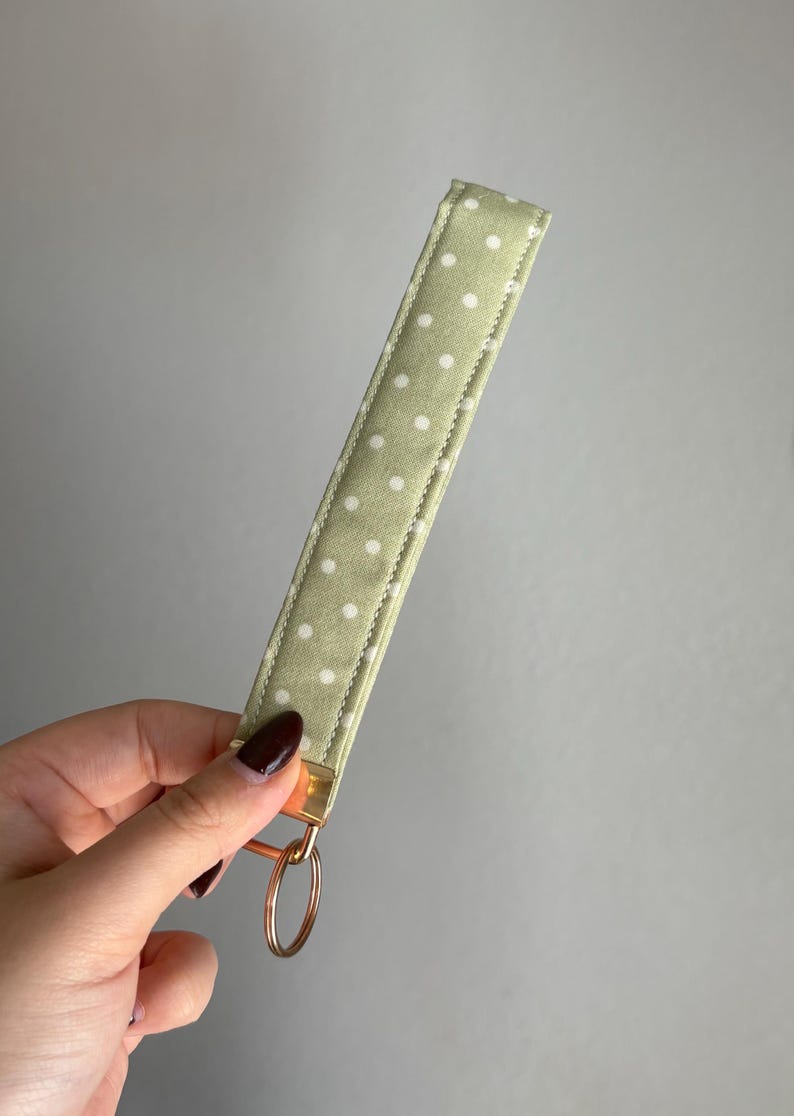 May include: A pale green key fob wrist strap with a white polka dot design. It features a gold-tone metal clasp and a copper-coloured key ring. The fabric has a soft, textured feel.