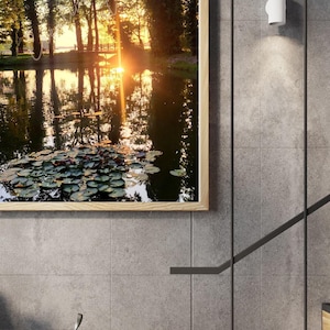 Puede incluir: Una impresión artística enmarcada que presenta una serena escena de estanque con nenúfares, árboles y una puesta de sol. La obra de arte se exhibe en una pared de azulejos grises, con una escalera moderna y una cesta de alambre.