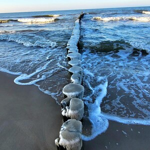 May include: A scenic coastal view featuring a line of weathered wooden posts extending into the ocean. The posts are covered in ice, with waves crashing around them. The sky is a clear blue.