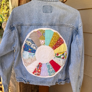 May include: A light blue denim jacket featuring a large, circular, multi-colored patchwork quilt design on the back. The circular patch consists of various floral and patterned fabric segments arranged around a solid white center circle.